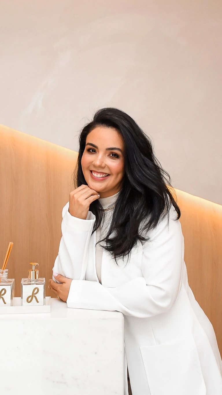 Adelaide Strube, cirurgiã-dentista, sorrindo com a mão no queixo, de jaleco branco, apoiada no balcão da recepção da Recriar.