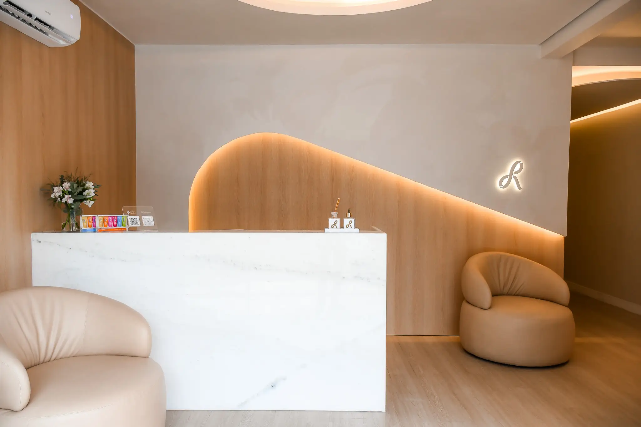 Recepção da Recriar Studio Odontológico: balcão de mármore claro, poltronas em tom areia e parede de madeira com iluminação âmbar em curva, sob o monograma R iluminado.