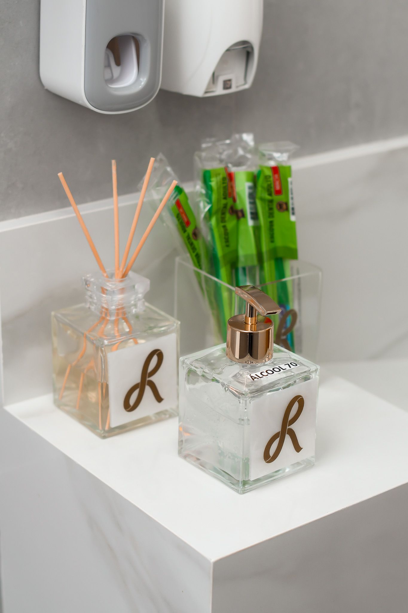 Bancada do lavabo com difusor, frasco de álcool em gel e escovas embaladas, sob dispensers de parede.